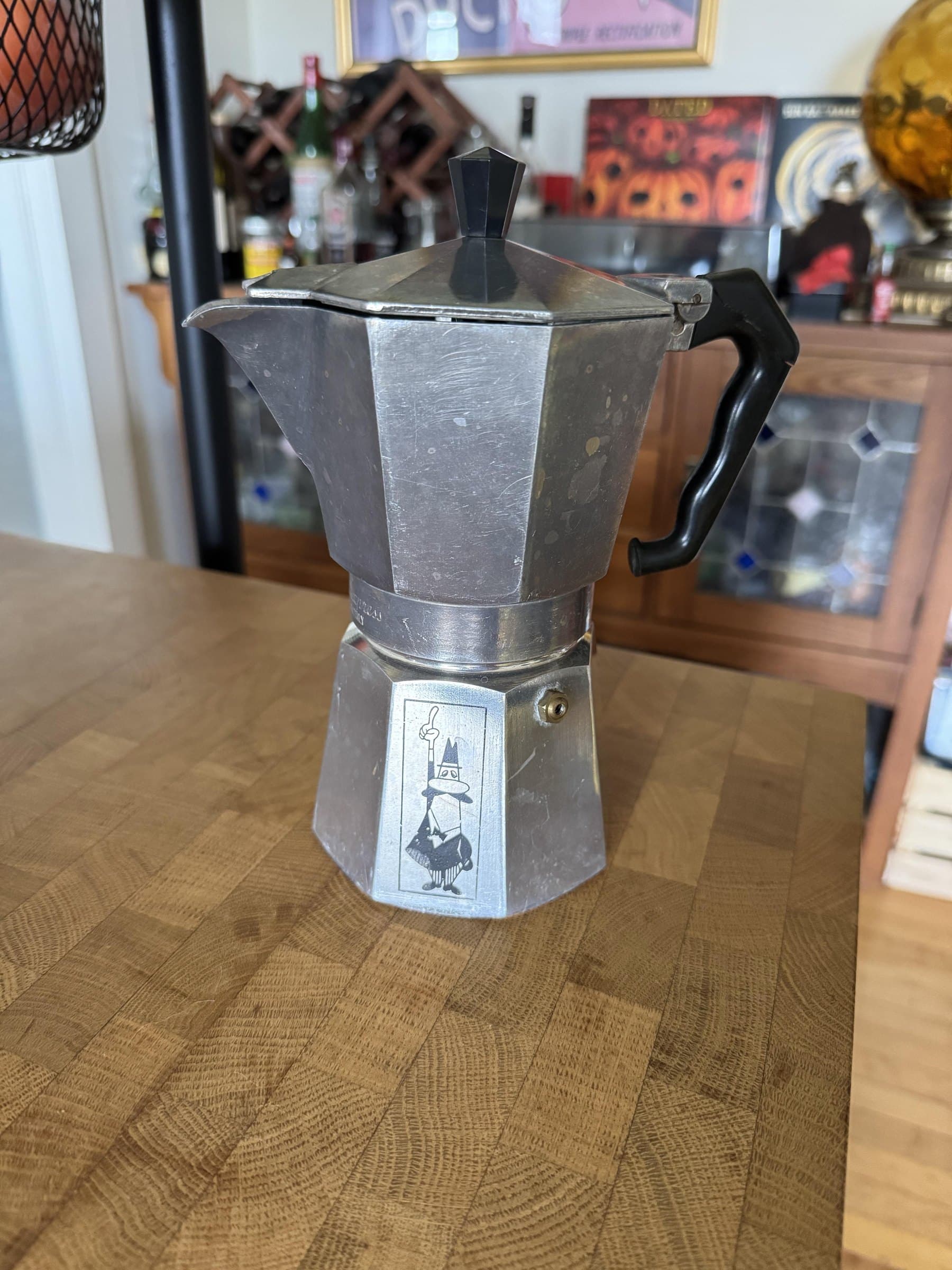 An old aluminium Moka Express on a butcher block counter, its finish scuffed and spotted, the Omino printed on the side of the base