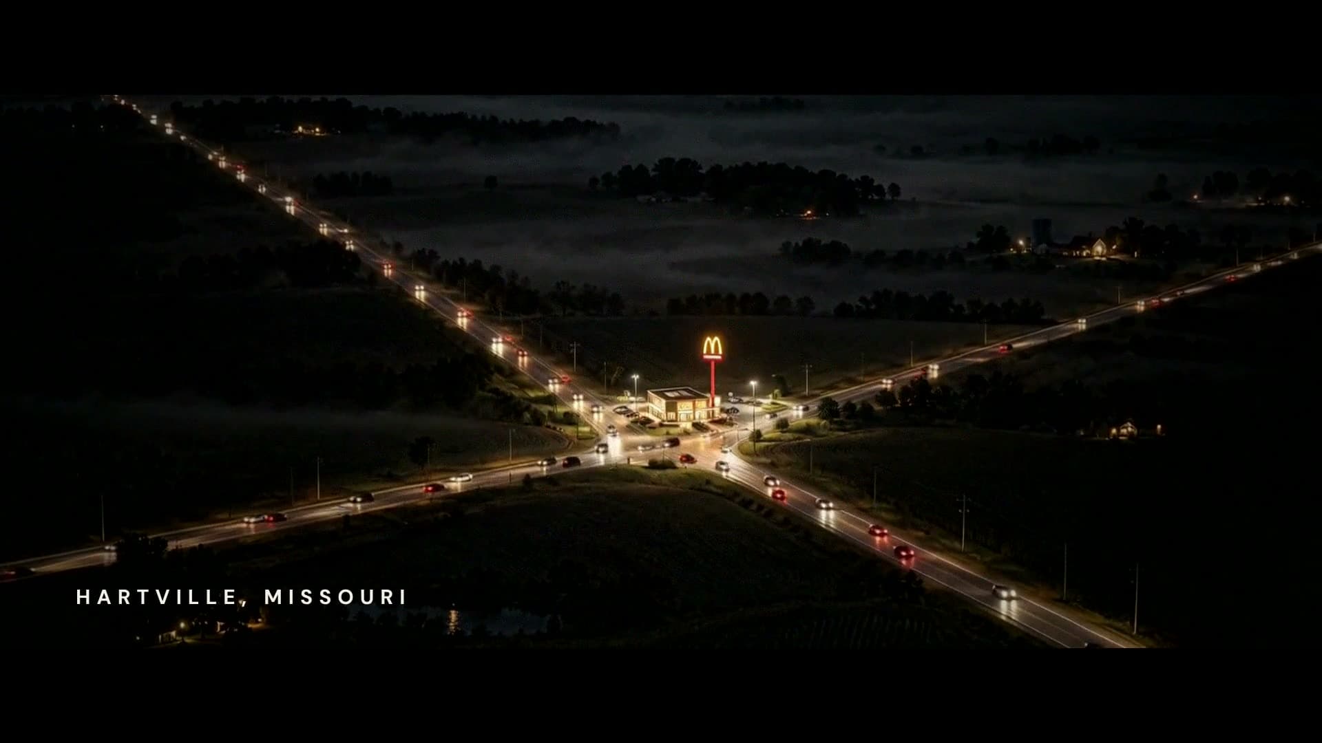 From the air at night, country roads cross at one lit McDonald's in a dark field, headlights streaming in from every side. The super reads Hartville, Missouri.