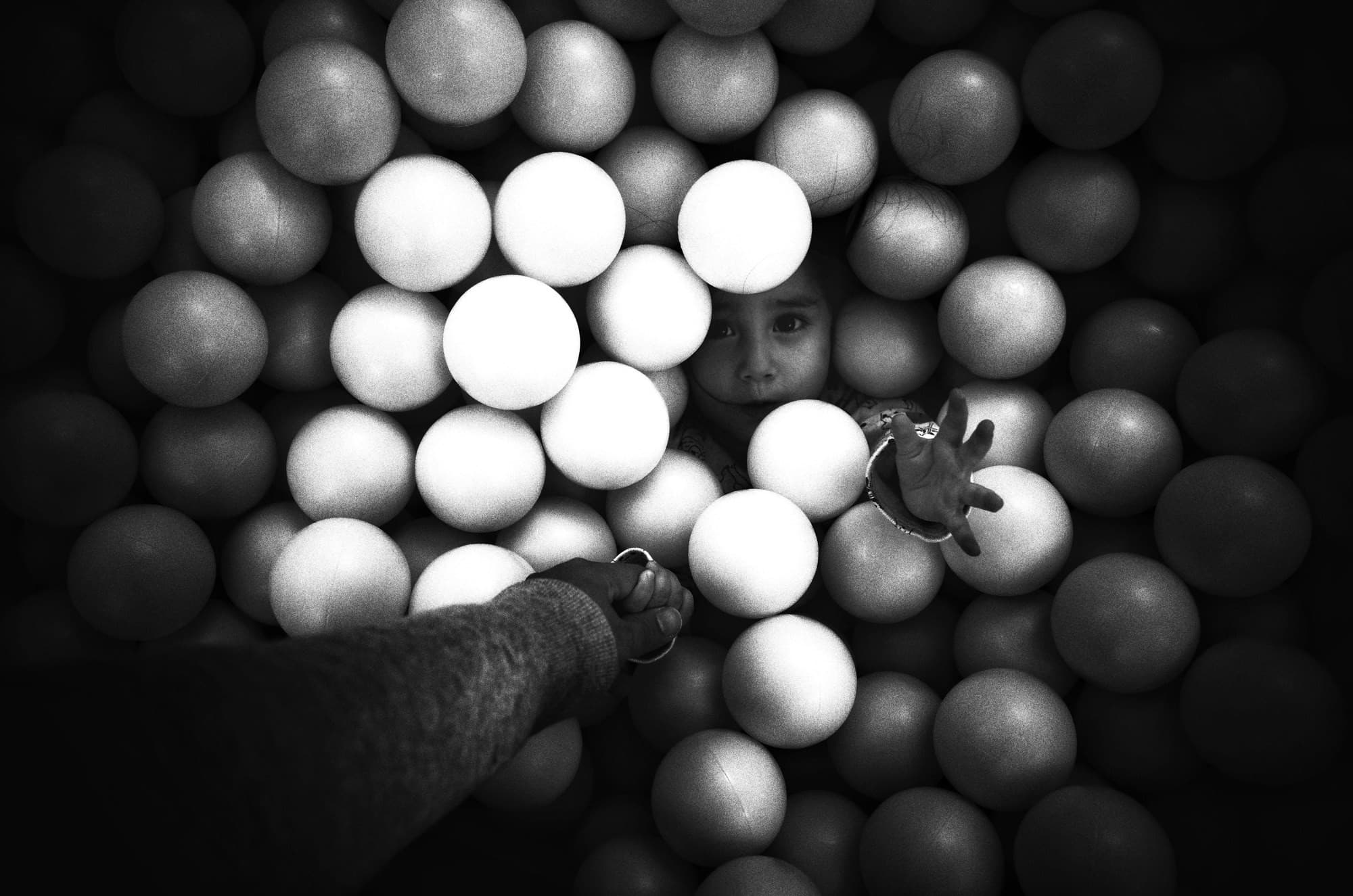 Black and white photo looking down into a ball pit. The photographer’s arm reaches in and holds the hand of a little girl peeking out between the balls