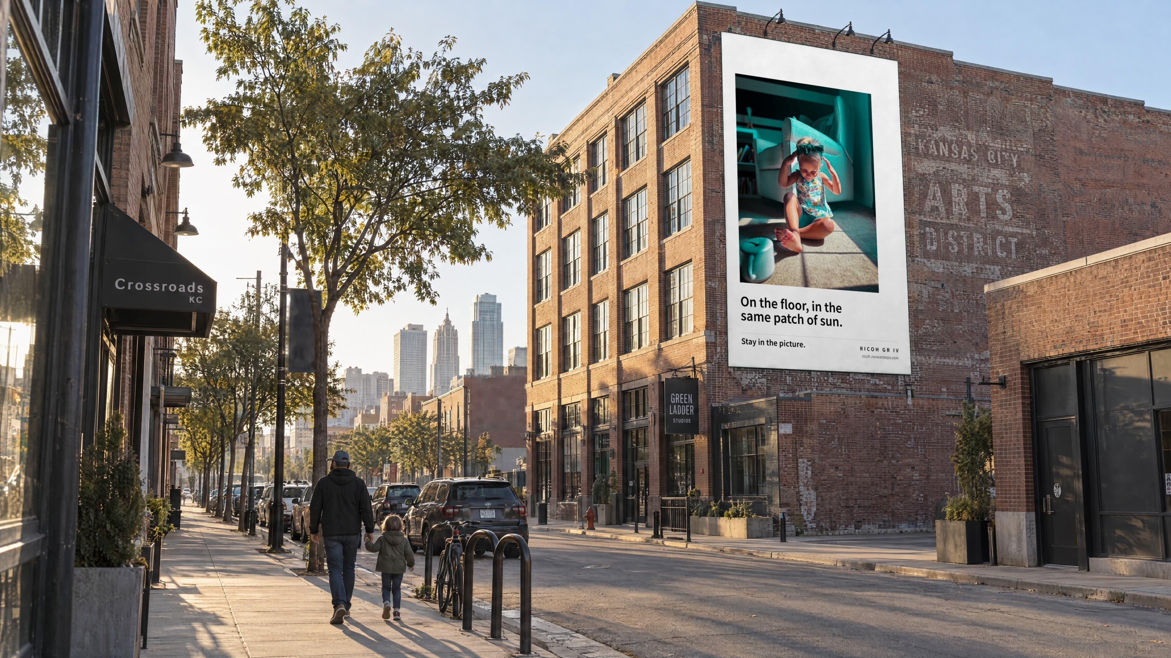 A wallscape on a brick building in the Kansas City Crossroads: a toddler in a patch of morning sun, with the line On the floor, in the same patch of sun. A parent and child walk hand in hand below.