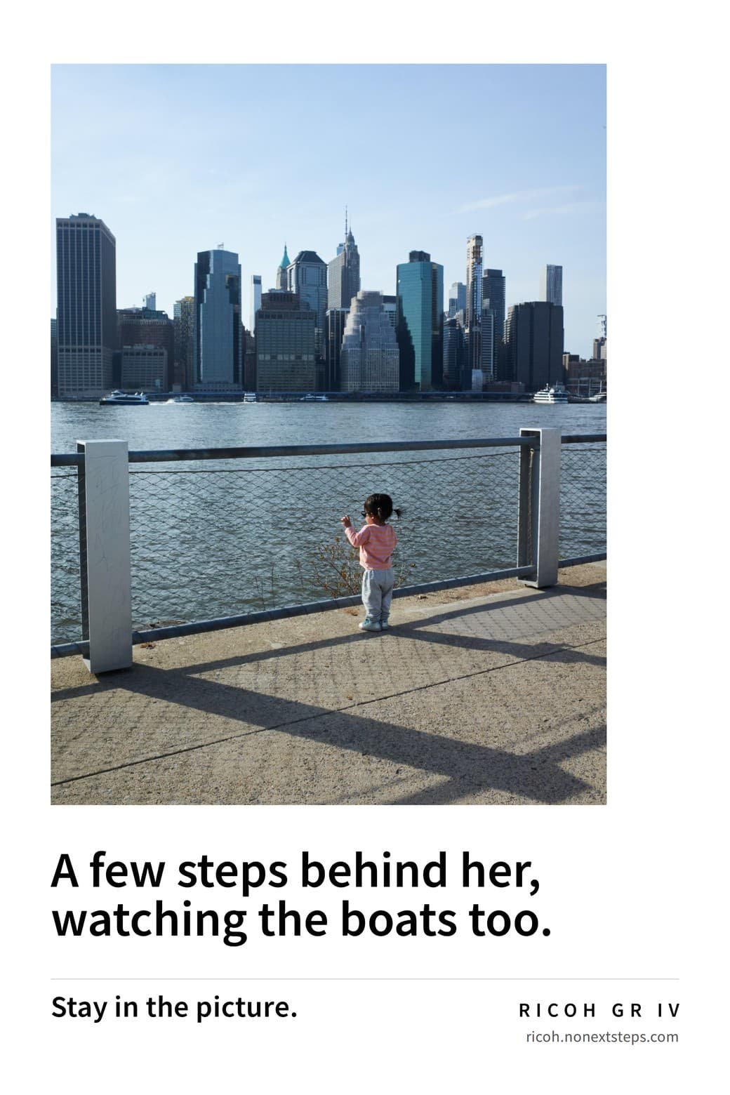 Poster: a small girl in pink at a river railing with the Manhattan skyline beyond. A few steps behind her, watching the boats too. Stay in the picture. RICOH GR IV